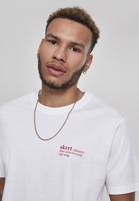 White cotton t-shirt featuring red text that reads "skrrt (skuur) that noise in every rap song." Accessorized with a thin gold chain.