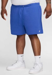 Blå bomullsshorts med elastisk midja och dragsko, med en vit Nike-logotyp på kanten. Bärs med vita sneakers och strumpor.