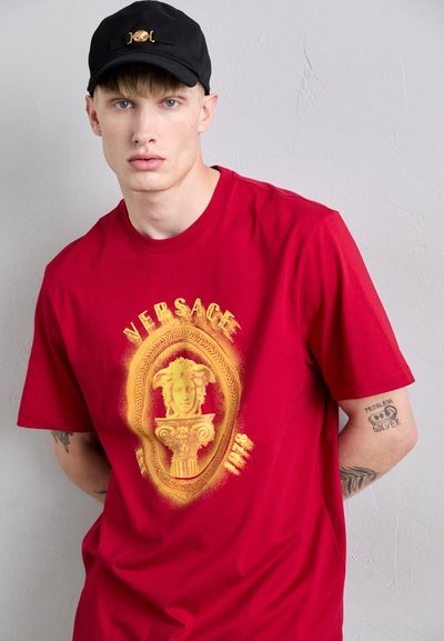 Jaunas vyras vilkintis raudonu Versace marškinėliu su Medūzos logotipu ir juoda kepure, demonstruojantis tatuiruotes ant dilbio, stovi prieš paprastą tekstūruotą sieną.