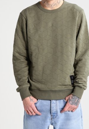 Mann trägt ein olivgrünes, gesteppte Sweatshirt und hellblaue Jeans, die Hände in den Taschen, mit sichtbaren Tätowierungen am Hals und an den Händen.