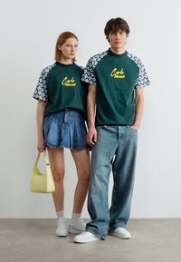 Νεαρή γυναίκα και άνδρας στέκονται δίπλα-δίπλα φορώντας ταιριαστά πράσινα μπλουζάκια με σχέδιο, τζιν παντελόνια, λευκά αθλητικά παπούτσια και ουδέτερες εκφράσεις.