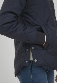 Henkilö, joka on pukeutunut tummansiniseen tikkitakkiin, jossa on nepparit, käsi sivutaskussa, yhdistettynä sinisiin farkkuihin ja valkoiseen hihaan, joka on näkyvissä.