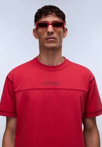 Κόκκινο πουκάμισο με κοντά μανίκια, στρογγυλή λαιμόκοψη και ραμμένες λεπτομέρειες· διαθέτει κεντημένο λογότυπο "NAPAPIJRI" στο στήθος.