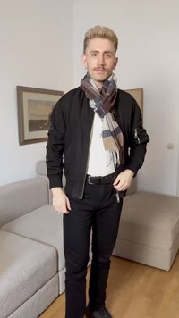 Veste bomber noire, chemise blanche, pantalon noir et écharpe à carreaux aux motifs gris, bleu et brun. Chaussures noires. Sol en bois.