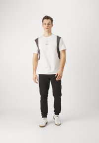 Witte t-shirt met donkergrijze schouderpanelen en een klein merklogo, gecombineerd met zwarte joggerbroeken en witte sneakers.