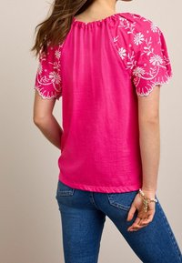 Rosa Oberteil mit gesticktem Blumenmuster auf den kurzen Ärmeln, ausgestattet mit einem gerafften Ausschnitt und glatter Textur, kombiniert mit blauen Jeans.
