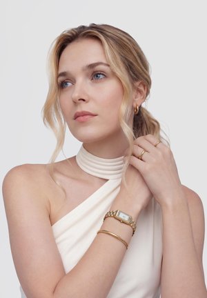 Femme aux cheveux blonds portant un haut licou blanc, une montre en or, un bracelet, une bague et des boucles d'oreilles créoles, regardant pensivement sur le côté.