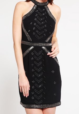 Robe noire sans manches à col licou avec des embellissements géométriques en perles argentées et une taille cintrée, portée par une femme le bras posé sur la hanche.