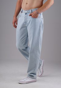 Oberkörperfreier Mann, der hellblaue Jeans und weiße Sneakers trägt, steht mit einer Hand in der Tasche vor einem schlichten grauen Hintergrund.