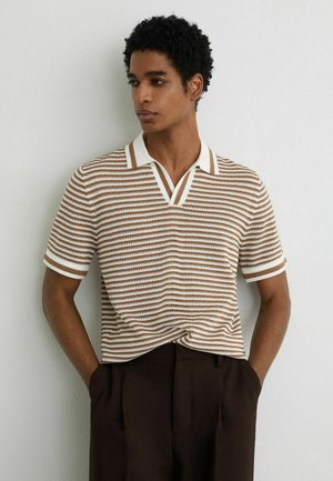 Göndör hajú férfi bézs és fehér csíkos rövid ujjú galléros pólóban és sötétbarna nadrágban, zsebében tartott kezekkel állva egy fehér fal előtt.