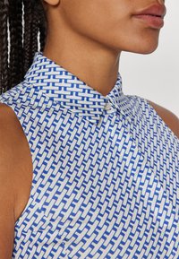 Collared sleeveless blouse featuring a geometric blue and white pattern. Made of smooth fabric with a button closure at the collar.