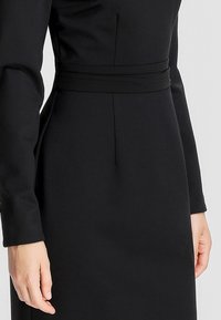 Gros plan sur une personne portant une robe noire ajustée à manches longues avec une couture horizontale à la taille, les mains reposant détendues le long du corps.