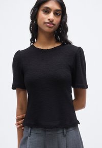 Μαύρο υφασμάτινο τοπ με κοντά μανίκια, με κυματοειδές ντεκολτέ και τελείωμα, που διαθέτει εφαρμοστή σιλουέτα και ελαφρώς φουσκωτά μανίκια.