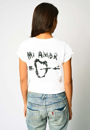 Kvinde med langt mørkt hår iført hvid t-shirt med sort tekst "Mi Amor" og hjerte gennemstikket af en pil, kombineret med lyseblå jeans.