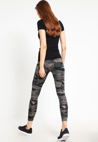 Donna con lunghi capelli rossi che indossa un top nero a maniche corte, leggings mimetici grigi e neri, e sneakers nere, in piedi su uno sfondo bianco.