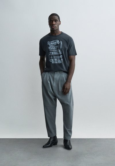 Άνδρας που φοράει σκούρο γραφικό μπλουζάκι με κείμενο, γκρι πιε-ντε-πουλ παντελόνι και μαύρες αστραγάλες μπότες, στέκεται μπροστά σε φωτεινό απλό φόντο.