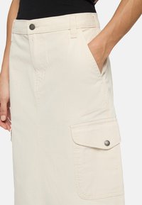 Beige cargokjol i bomull, med en sidoficka med lock och tryckknapp, bältesöglor och synliga söms detaljer.