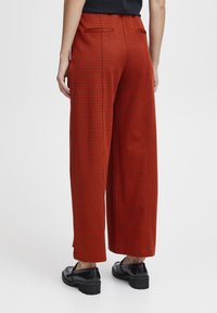 Pantalons larges à carreaux rouges avec deux poches arrière, une texture lisse, associés à des chaussures noires brillantes sur un fond uni.