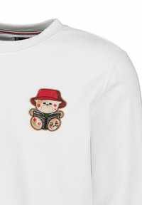 Weißer Pullover mit einem gestickten Bären, der einen roten Hut trägt, ein Buch hält und dekorative Symbole in roten und braunen Akzenten zeigt.