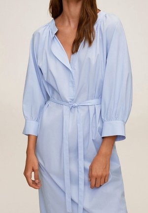 Vaaleansininen mekko, joka on valmistettu sileästä kankaasta. Siinä on klassinen kaulus, pitkät halkiopuiset hihat ja vyötärönauha säädettävää istuvuutta varten.