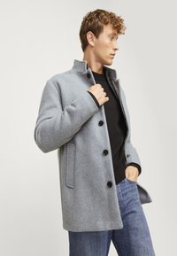 Abrigo de mezcla de lana gris con un cuello alto, cuatro botones negros y bolsillos laterales, combinado con un suéter negro y jeans azules.