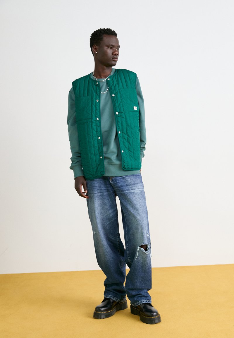 Zelená prošívaná vesta přes teplákovou mikinu v barvě teal, doplněná o oprané modré džíny a černé platformové boty. Stojící na žluté podlaze.