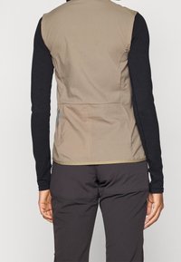 Beżowa kamizelka bez rękawów z lekkiego materiału, z kieszenią na boku i gładką teksturą. Połączona z czarnymi długimi rękawami i ciemnymi spodniami.