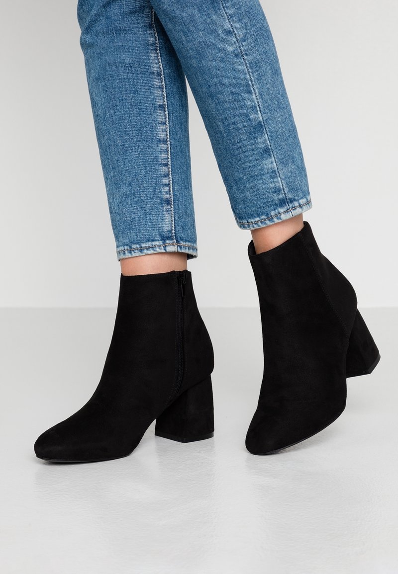 Personne portant un jean bleu et des bottines noires en suède à talons blocs, debout sur un sol clair.
