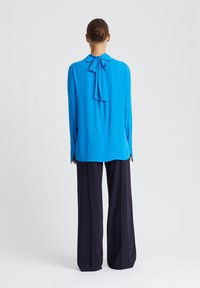 Langärmlige blaue Bluse mit einer Schleife am Kragen, glatter Textur und geknöpften Manschetten, kombiniert mit hoch taillierten, dunklen, weiten Hosen.