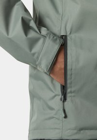 Chaqueta impermeable verde que presenta un primer plano de un bolsillo con cremallera y puños ajustables, mostrando una textura suave y mate.