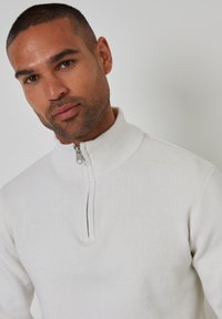 Weißer Reißverschluss-Pullover mit hohem Rippkragen, enganliegendem Design, glatter Textur und kontrastierendem Reißverschlussdetail am Hals.