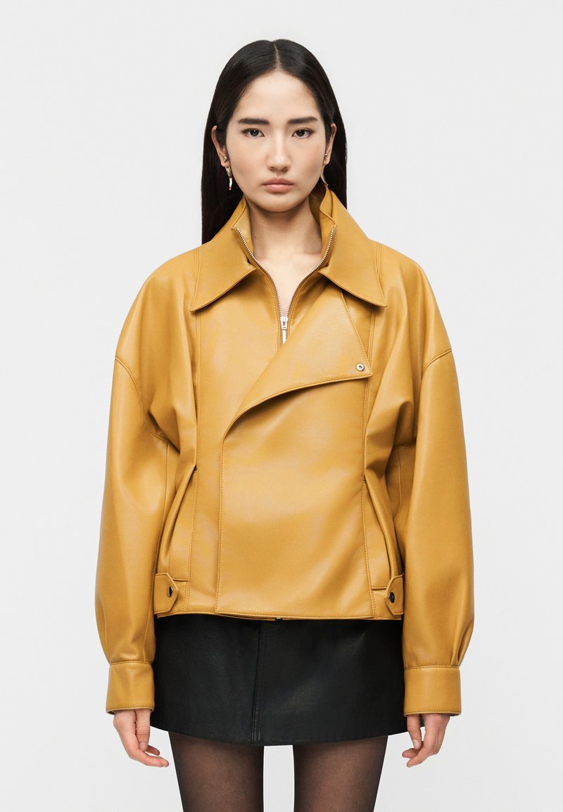 Γυναίκα με μακριά σκούρα μαλλιά φοράει υπερμεγέθη δερμάτινο μπουφάν μουσταρδί και μαύρη μίνι φούστα σε λευκό φόντο.