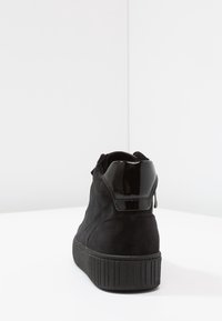 Black suede sneaker with a high platform sole and glossy black heel detail, shown from the back on a white surface.