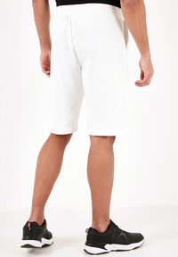Hvite bomullsshorts med elastisk talje, der når midt på låret. Personen bærer sorte sportsko med hvite såler.