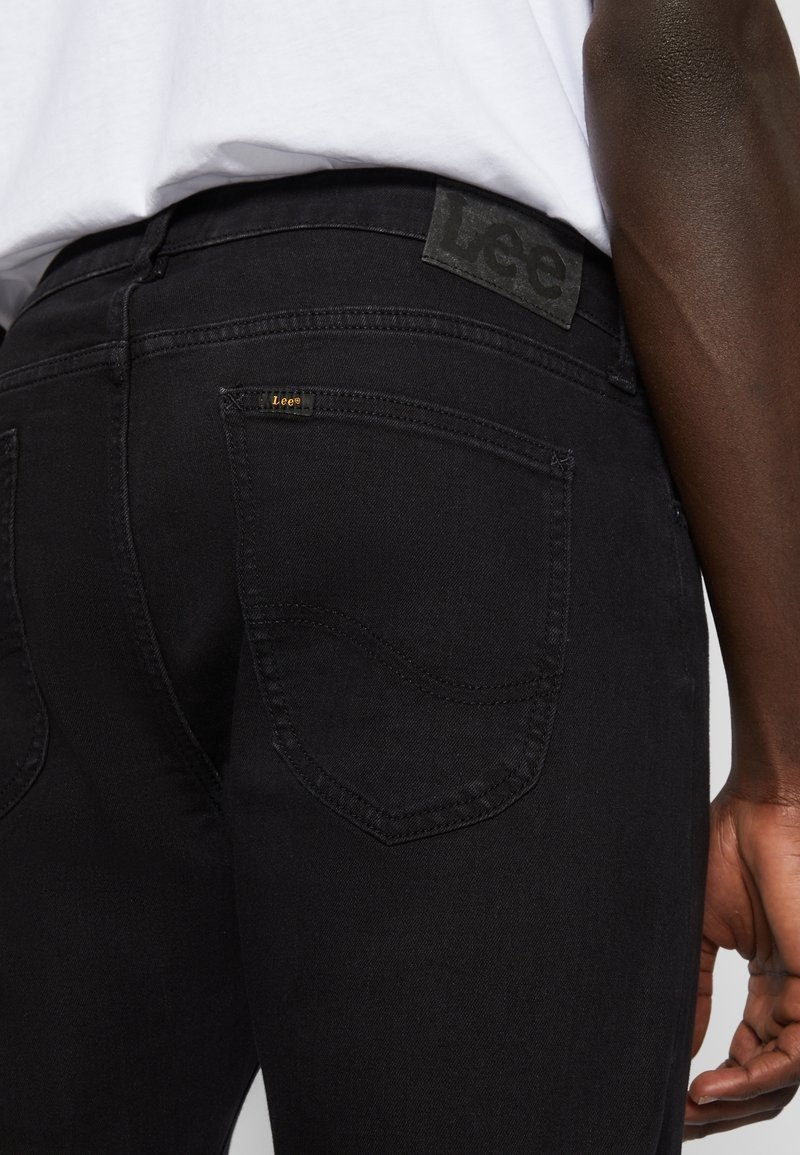 Gros plan d’une personne portant un jean Lee noir avec une poche arrière visible et un écusson logo sur la ceinture, chemise blanche rentrée.