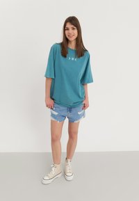 T-shirt oversize turchese con grafica di luna e stella, abbinata a pantaloni corti in denim rasato e sneakers alte beige.