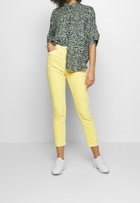 Žena v žlutých zkrácených kalhotách, s volnou košilí s hadím vzorem, bílými teniskami, stojící s jednou rukou v kapse na jednobarevném pozadí.