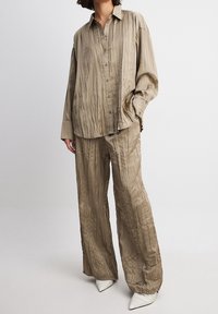 Beżowy, pomarszczony zestaw koszuli i spodni charakteryzuje się luźnym krojem, guzikiem z przodu oraz długimi rękawami. Połączony z białymi szpilkami z ostrym noskiem.