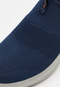 Scarpa slip-on lavorata a maglia blu con tomaia testurizzata, design elastico e suola in gomma bianca a contrasto. Presenta un sottile accento con logo arancione.