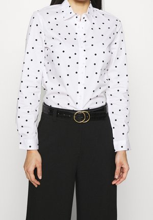 Femme portant une chemise blanche à manches longues à boutons avec des pois noirs, un pantalon noir et une ceinture noire avec des doubles anneaux dorés.