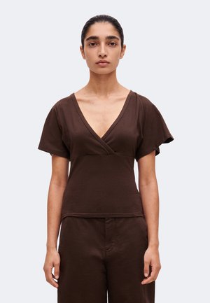 Femme aux cheveux foncés tirés en arrière, portant un haut marron à manches courtes avec un profond décolleté en V et un pantalon marron assorti sur un fond clair uni.