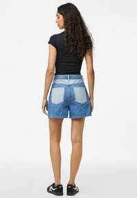 Augstās vidukļa zila džinsu šorti ar brīvu piegriezumu, ar divām aizmugurējām kabatām un neapstrādātu maliņu. Apvienoti ar melnu T-kreklu un sporta apaviem.