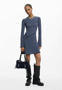 Robe longue à manches longues côtelée bleue avec ourlet asymétrique, associée à un sac à bandoulière noir texturé et des bottes noires brillantes montant jusqu'aux genoux.