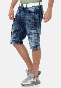 Dunkelblaue Denim-Cargo-Shorts mit mehreren Reißverschlusstaschen und strukturierten Details. Kombiniert mit grauen Sportschuhen.