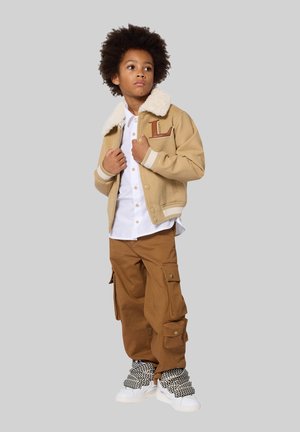 Braune Bomberjacke mit weißem Kunstpelzkragen, braune Cargo-Hose, weißes Hemd und schwarz-weiße Sneaker mit Muster. Lässiges Outfit.