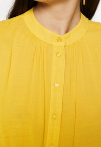 Blusa gialla con bottoni, caratterizzata da un collo tondo, una texture plissettata e un tessuto opaco. I bottoni sono coordinati per colore e di forma rotonda.