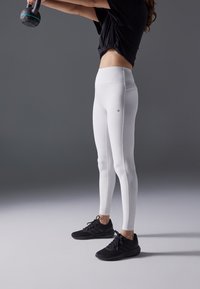 Valkoiset legginsit, joissa on sileä pinta ja korkea vyötärö. Yhdistetty mustiin urheilujalkineisiin. Ei näkyviä kuvioita tai varusteita.