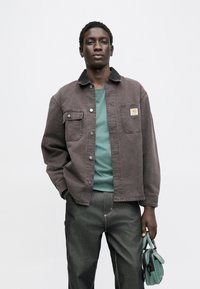 Hnědá džínová bunda s černým límcem, dvěma předními kapsami a stříbrnými knoflíky přes tyrkysové tričko, doplněná tmavými džíny a zelenou kabelkou.