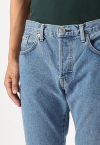 Detailní záběr na osobu ve světlých modrých džínech a tmavě zeleném tričku zastrčeném v pase, s jednou rukou uvolněně položenou vedle těla.