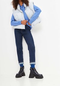 Gilet imbottito azzurro chiaro sopra una camicia a maniche lunghe blu, abbinato a jeans scuri in denim e stivali neri chunky con lacci.